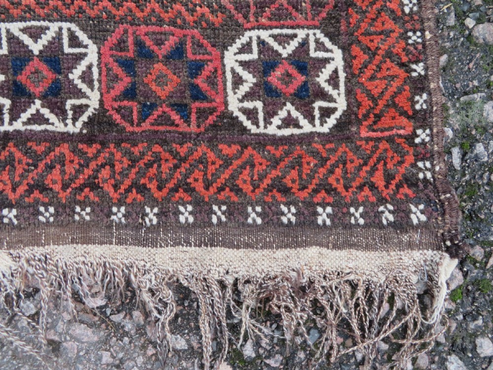 An antique Baluch wool rug featuring a repeating grid pattern of geometric motifs in rust red, dark blue, brown, and ivory accents with a fringed border. View IX