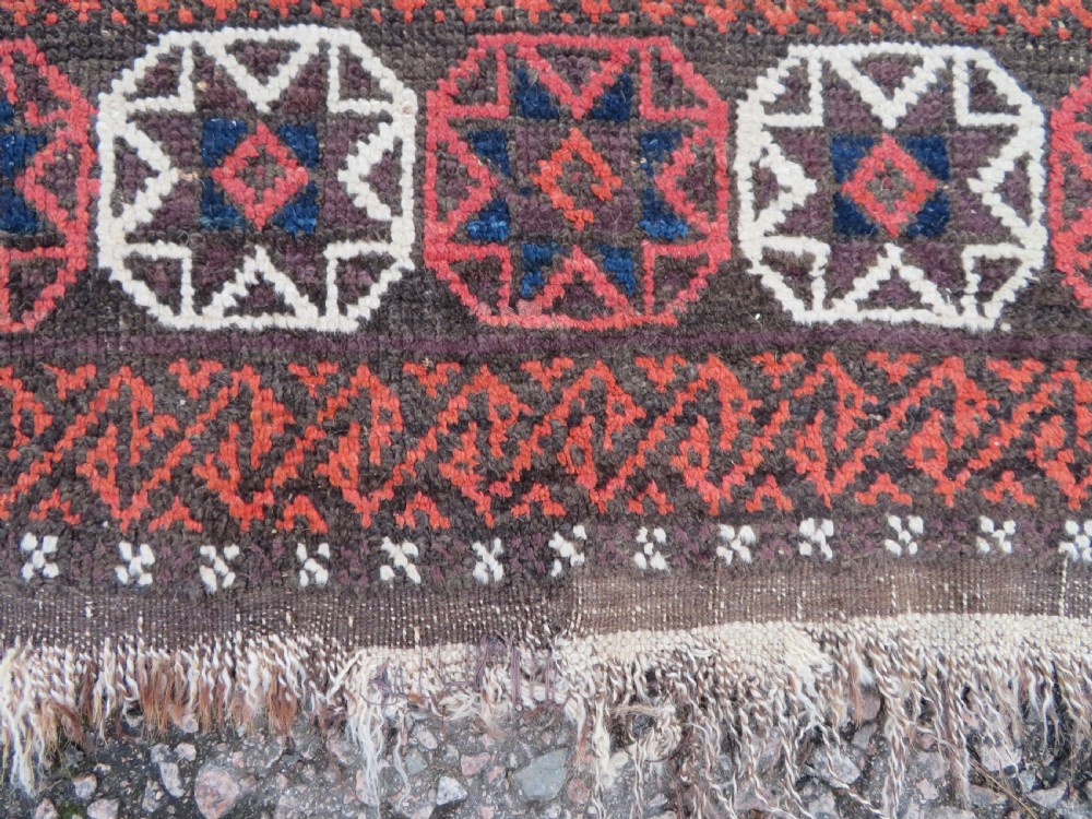 An antique Baluch wool rug featuring a repeating grid pattern of geometric motifs in rust red, dark blue, brown, and ivory accents with a fringed border. View X