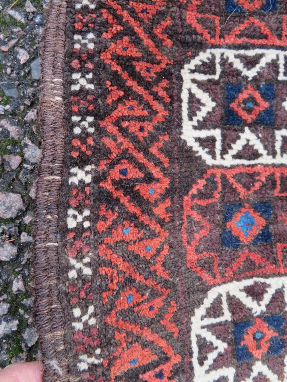 An antique Baluch wool rug featuring a repeating grid pattern of geometric motifs in rust red, dark blue, brown, and ivory accents with a fringed border. View XI