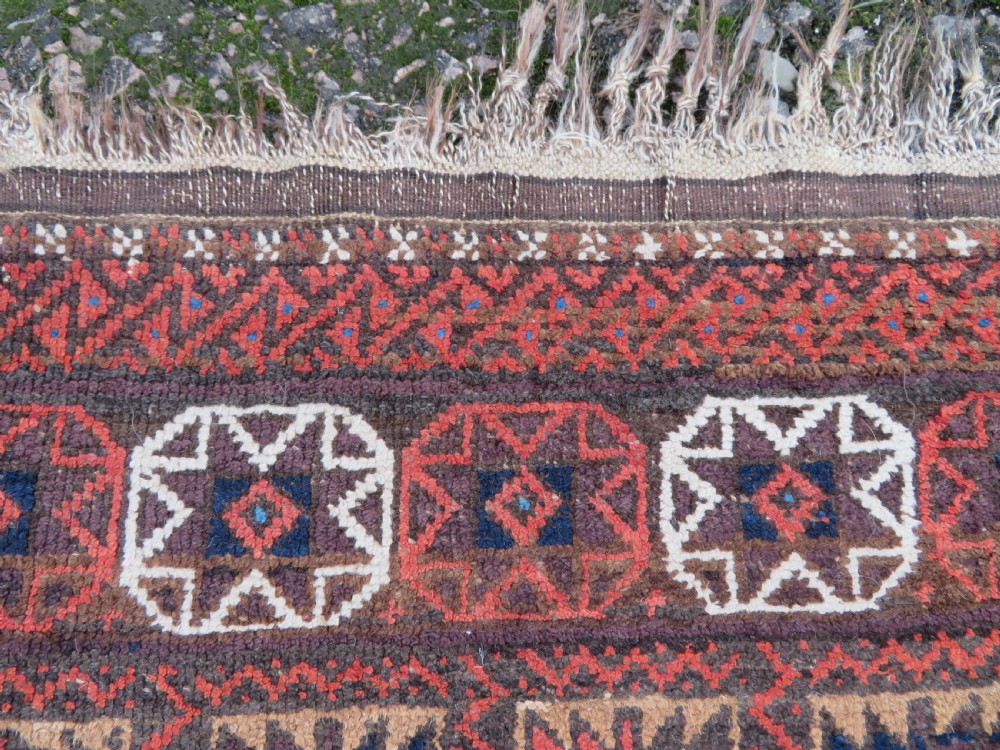 An antique Baluch wool rug featuring a repeating grid pattern of geometric motifs in rust red, dark blue, brown, and ivory accents with a fringed border. View XII