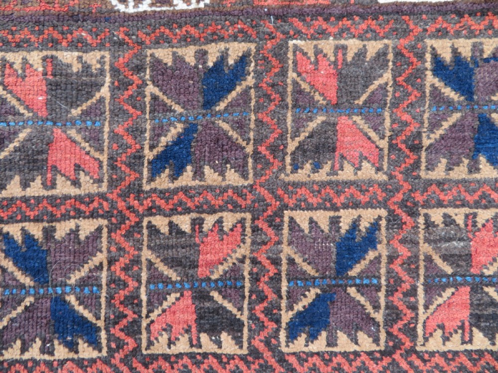 An antique Baluch wool rug featuring a repeating grid pattern of geometric motifs in rust red, dark blue, brown, and ivory accents with a fringed border. View XIII