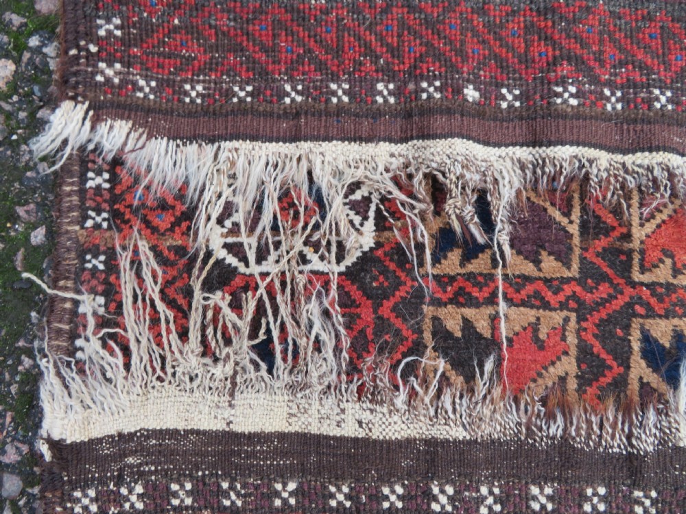 An antique Baluch wool rug featuring a repeating grid pattern of geometric motifs in rust red, dark blue, brown, and ivory accents with a fringed border. View XV