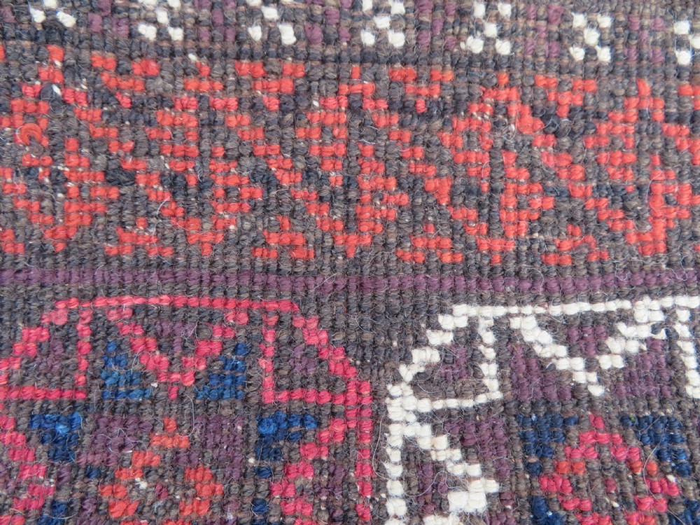 An antique Baluch wool rug featuring a repeating grid pattern of geometric motifs in rust red, dark blue, brown, and ivory accents with a fringed border. View XVI