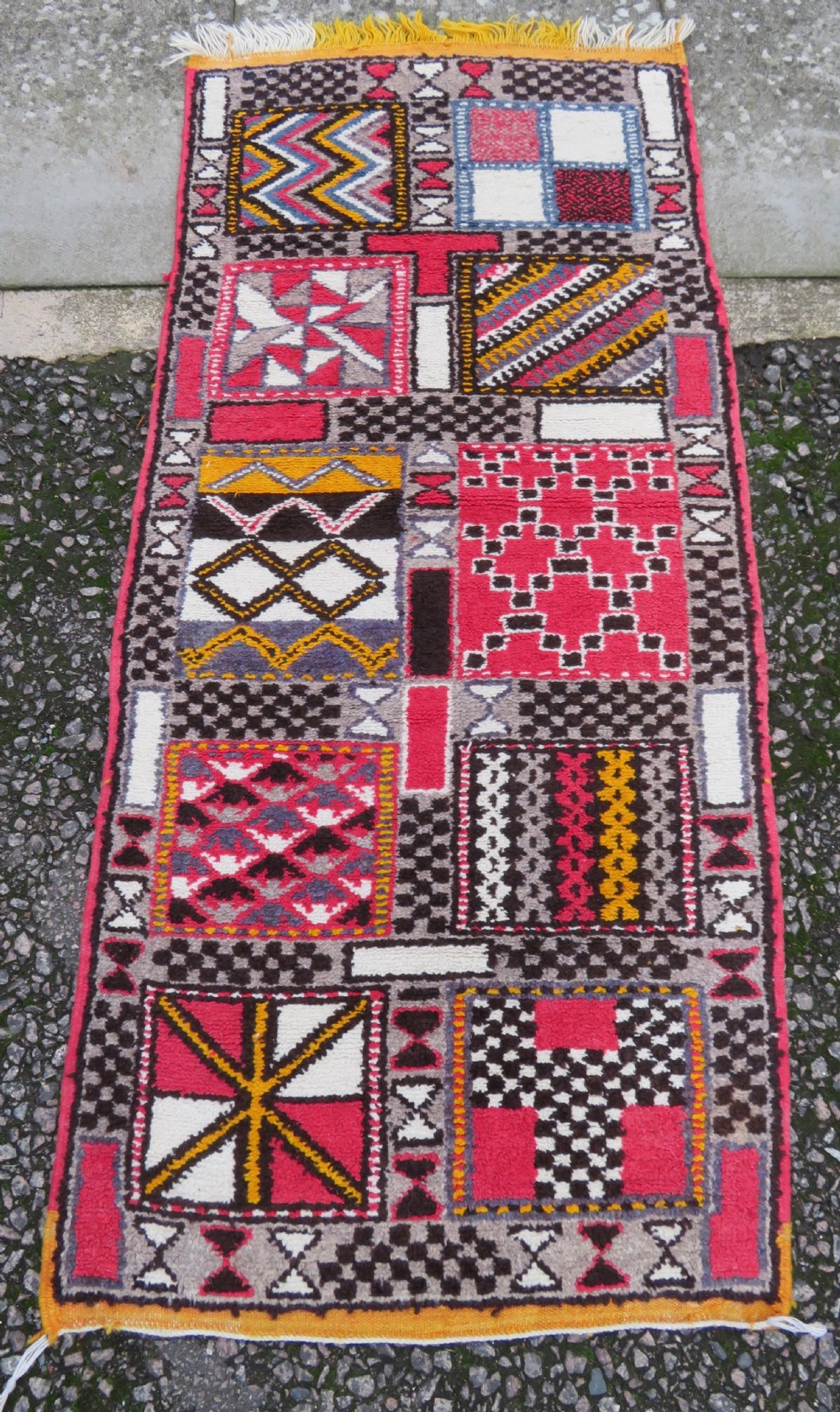 A rectangular hand-knotted Moroccan Berber wool rug featuring geometric patchwork panels in pink, yellow, black, grey, and white with fringed ends.