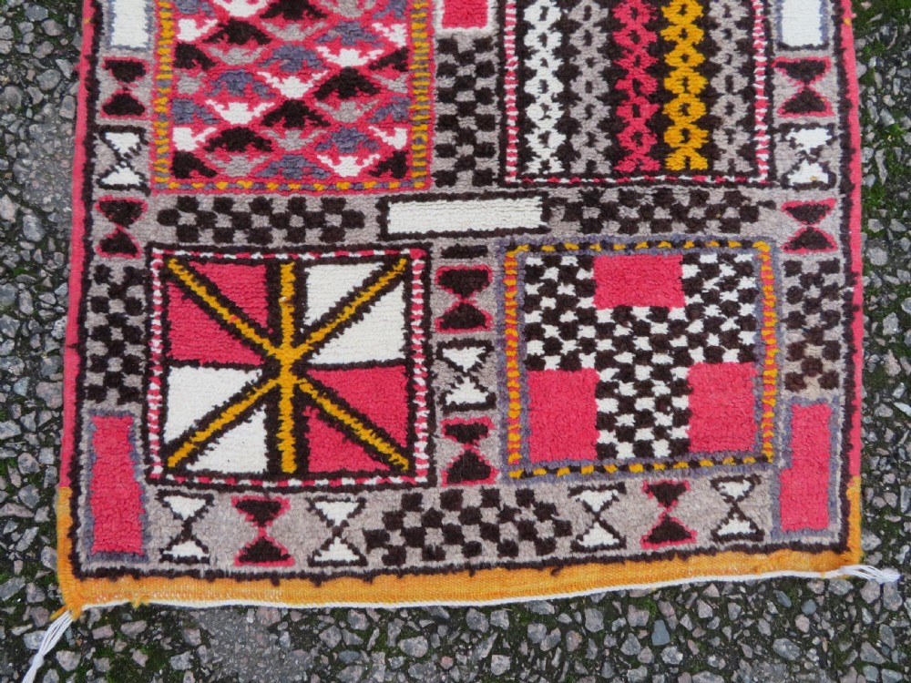 A rectangular hand-knotted Moroccan Berber wool rug featuring geometric patchwork panels in pink, yellow, black, grey, and white with fringed ends. View VI