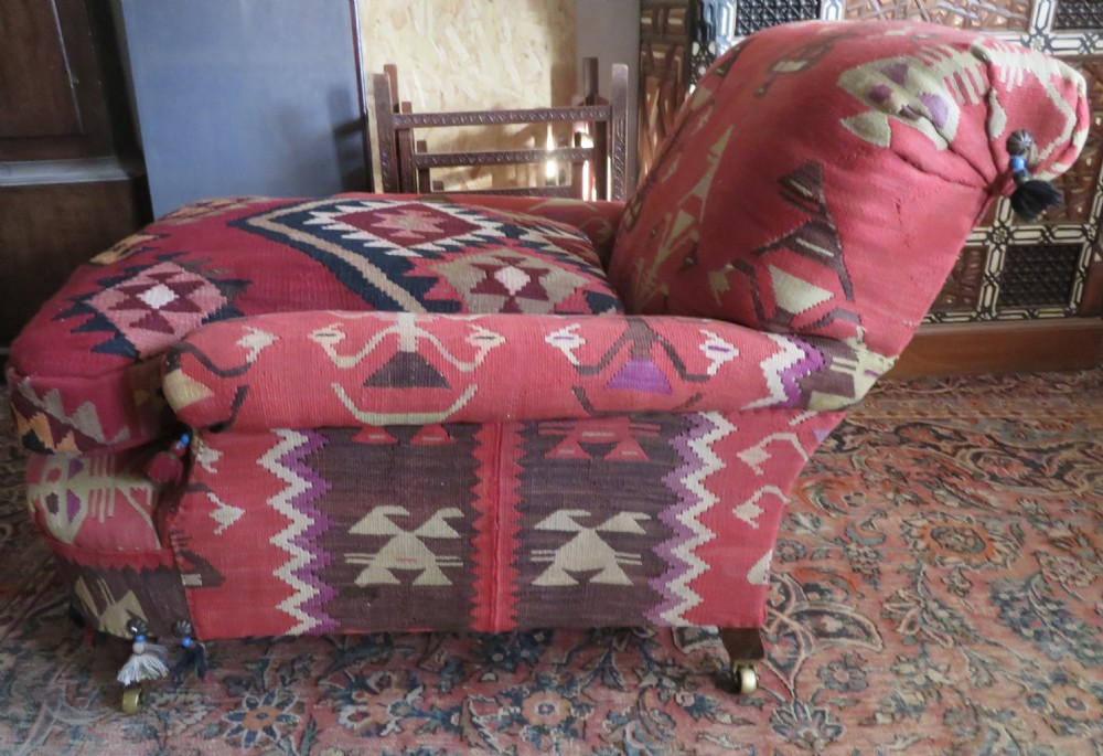 An antique low-slung club armchair upholstered in a geometric red wool kilim textile with turned legs and brass castors. View III