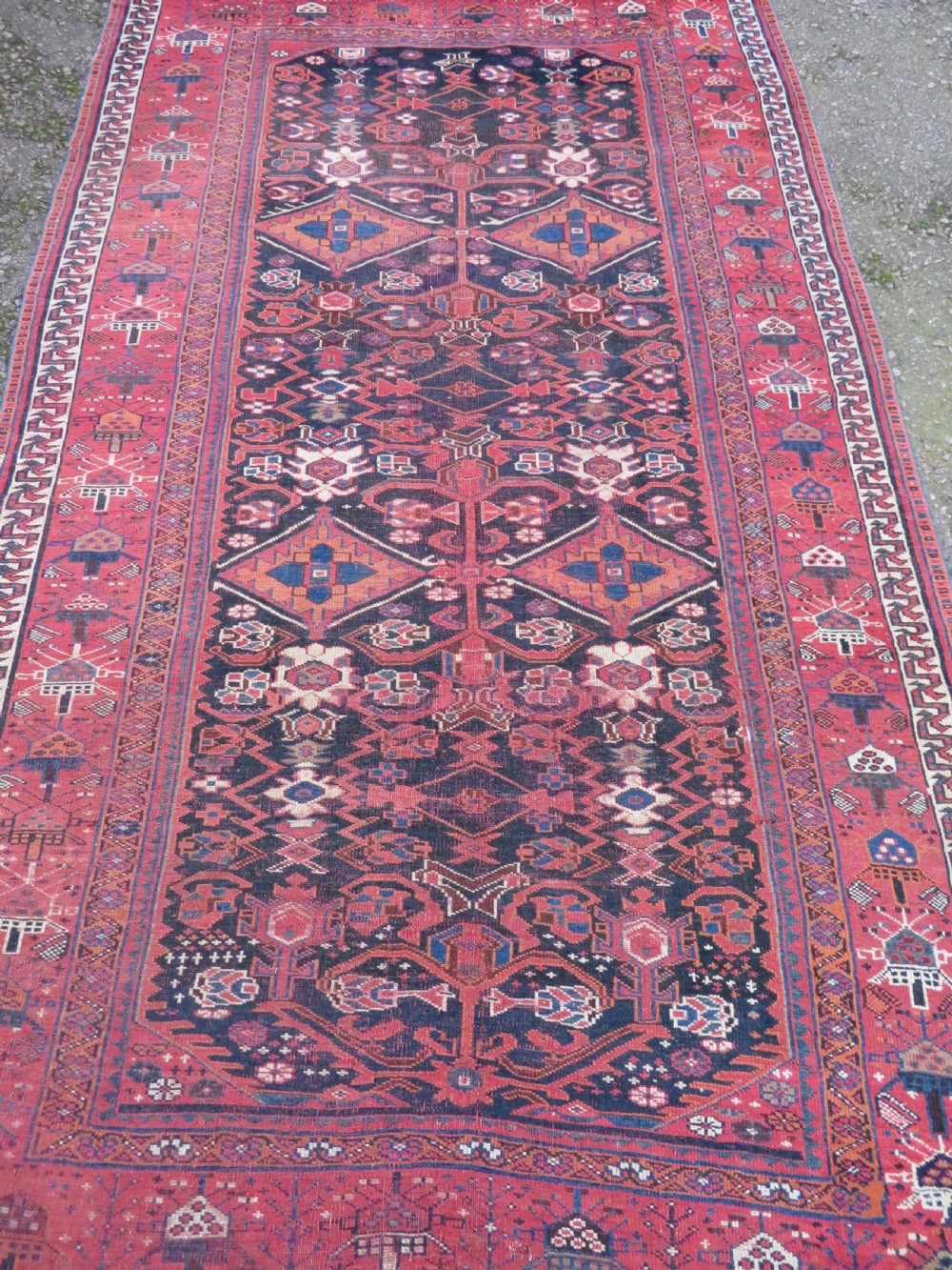 A large hand-knotted Persian Bakhtiari rug featuring geometric diamond medallions on a dark navy field surrounded by a detailed red patterned border. View IV