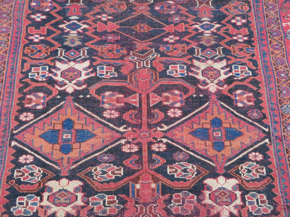 A large hand-knotted Persian Bakhtiari rug featuring geometric diamond medallions on a dark navy field surrounded by a detailed red patterned border. View V