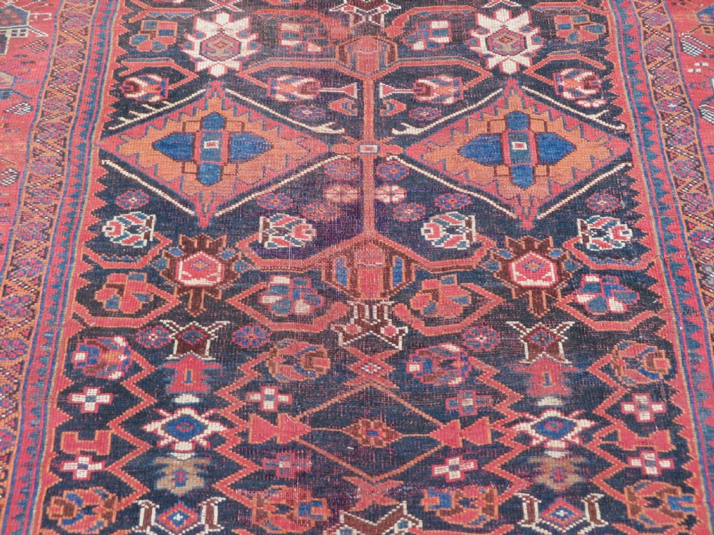 A large hand-knotted Persian Bakhtiari rug featuring geometric diamond medallions on a dark navy field surrounded by a detailed red patterned border. View VI