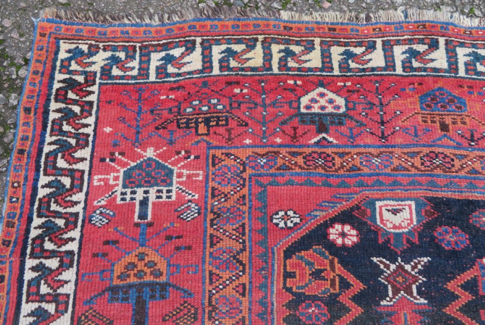 A large hand-knotted Persian Bakhtiari rug featuring geometric diamond medallions on a dark navy field surrounded by a detailed red patterned border. View VII