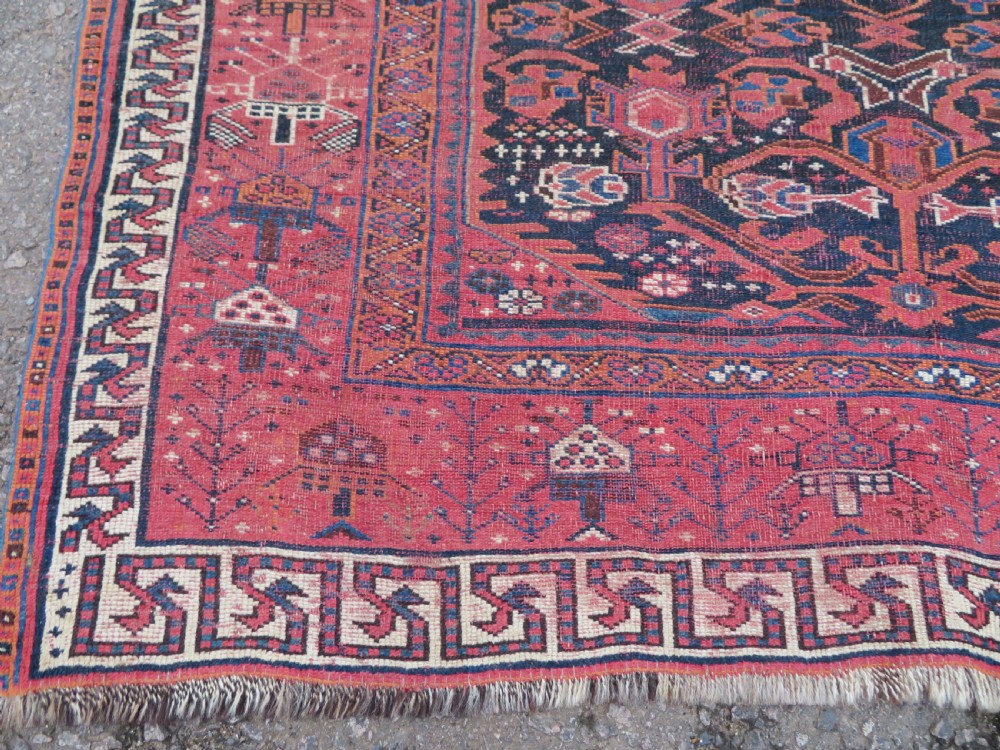 A large hand-knotted Persian Bakhtiari rug featuring geometric diamond medallions on a dark navy field surrounded by a detailed red patterned border. View X