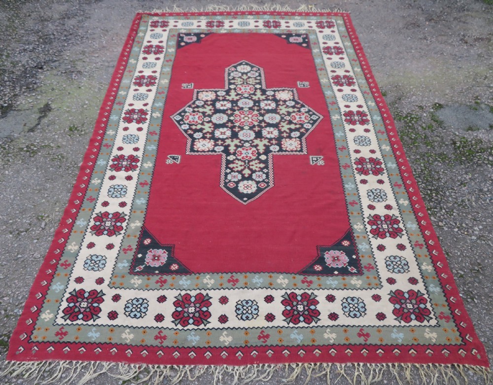 An antique flatweave Bessarabian wool rug featuring a red field with a central black and cream floral medallion, surrounded by a wide floral border and fringed edges.