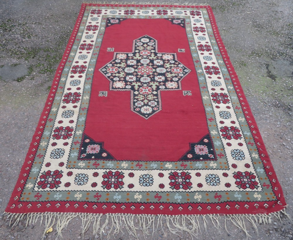 An antique flatweave Bessarabian wool rug featuring a red field with a central black and cream floral medallion, surrounded by a wide floral border and fringed edges. View II