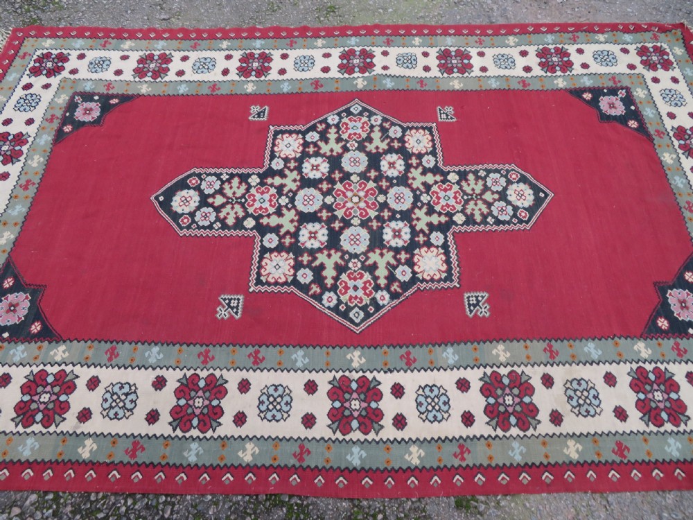 An antique flatweave Bessarabian wool rug featuring a red field with a central black and cream floral medallion, surrounded by a wide floral border and fringed edges. View III