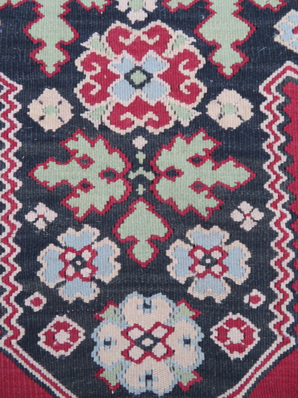 An antique flatweave Bessarabian wool rug featuring a red field with a central black and cream floral medallion, surrounded by a wide floral border and fringed edges. View V