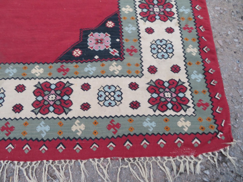 An antique flatweave Bessarabian wool rug featuring a red field with a central black and cream floral medallion, surrounded by a wide floral border and fringed edges. View VII