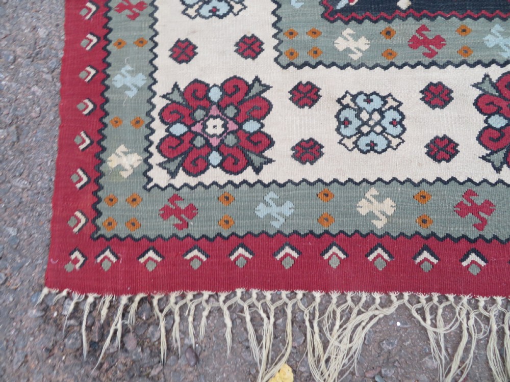 An antique flatweave Bessarabian wool rug featuring a red field with a central black and cream floral medallion, surrounded by a wide floral border and fringed edges. View IX