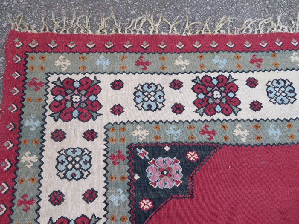 An antique flatweave Bessarabian wool rug featuring a red field with a central black and cream floral medallion, surrounded by a wide floral border and fringed edges. View XI
