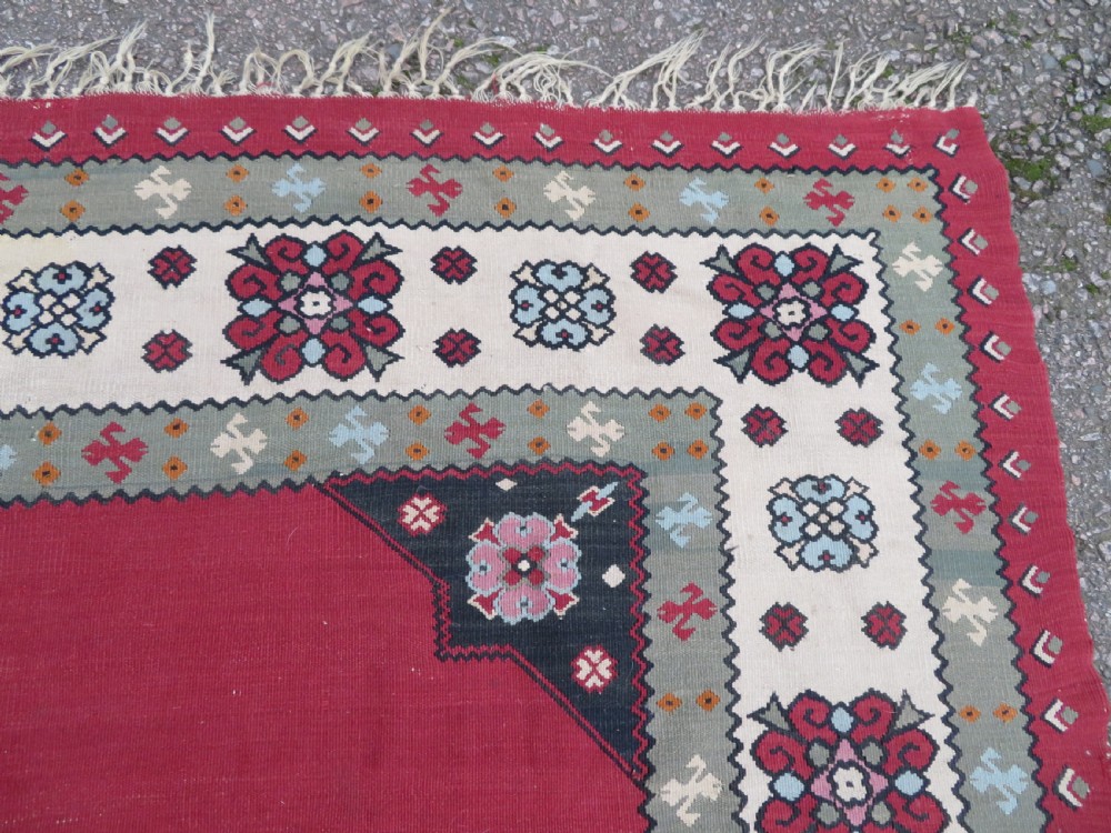 An antique flatweave Bessarabian wool rug featuring a red field with a central black and cream floral medallion, surrounded by a wide floral border and fringed edges. View XII