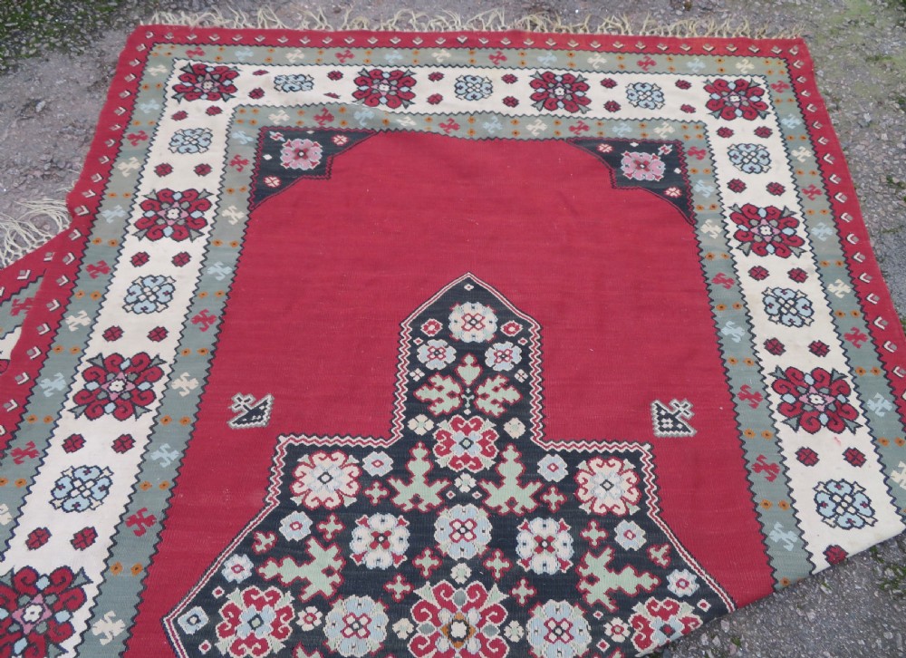 An antique flatweave Bessarabian wool rug featuring a red field with a central black and cream floral medallion, surrounded by a wide floral border and fringed edges. View XVII