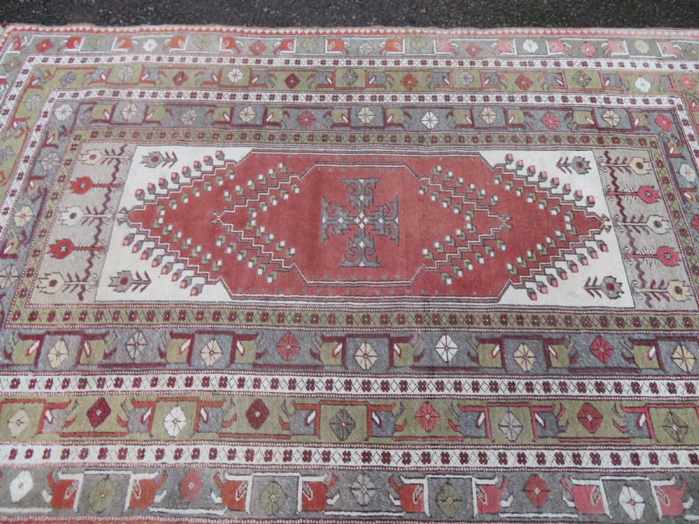 A hand-knotted antique Turkish Melas wool carpet featuring a terracotta red geometric diamond medallion on an ivory field with complex geometric borders. View IV