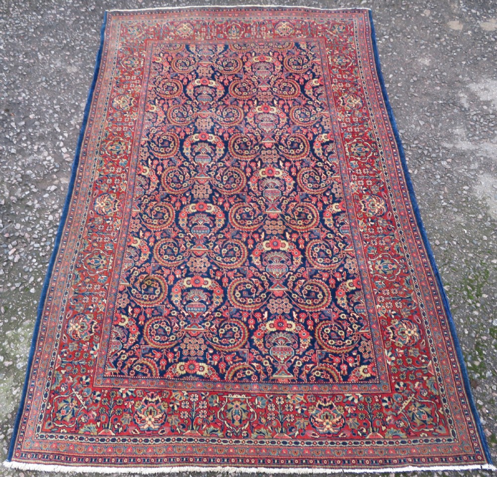 An antique hand-knotted Persian Kashan rug featuring an intricate blue central field with repeating floral urn motifs surrounded by a detailed red multi-banded border.