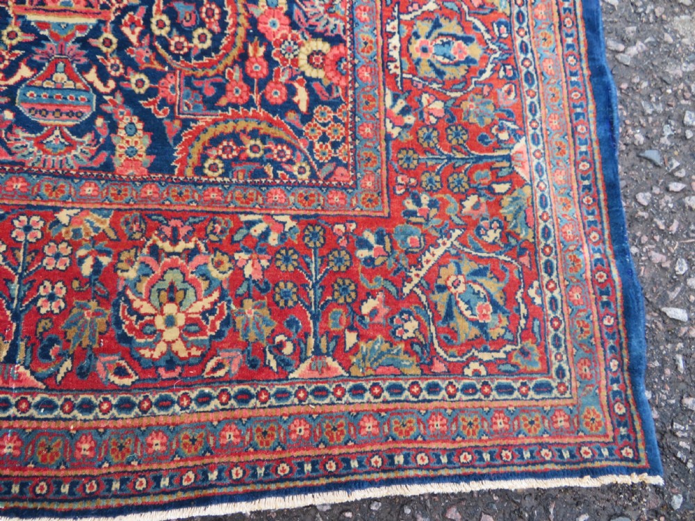 An antique hand-knotted Persian Kashan rug featuring an intricate blue central field with repeating floral urn motifs surrounded by a detailed red multi-banded border. View VI
