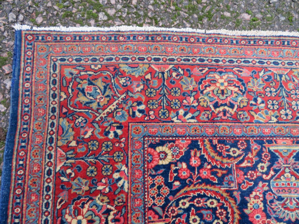 An antique hand-knotted Persian Kashan rug featuring an intricate blue central field with repeating floral urn motifs surrounded by a detailed red multi-banded border. View VII
