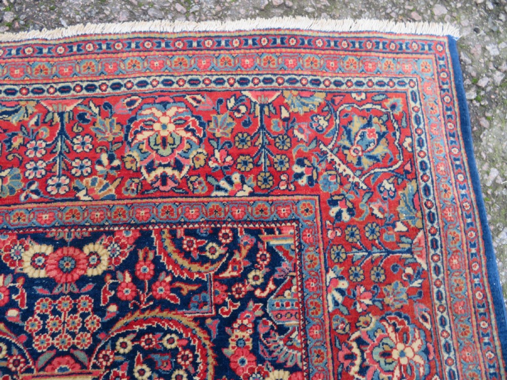 An antique hand-knotted Persian Kashan rug featuring an intricate blue central field with repeating floral urn motifs surrounded by a detailed red multi-banded border. View VIII