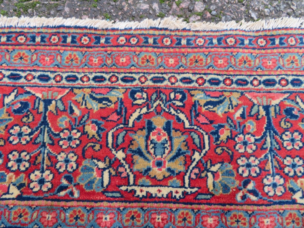 An antique hand-knotted Persian Kashan rug featuring an intricate blue central field with repeating floral urn motifs surrounded by a detailed red multi-banded border. View X