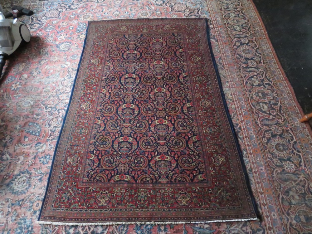 An antique hand-knotted Persian Kashan rug featuring an intricate blue central field with repeating floral urn motifs surrounded by a detailed red multi-banded border. View XVI