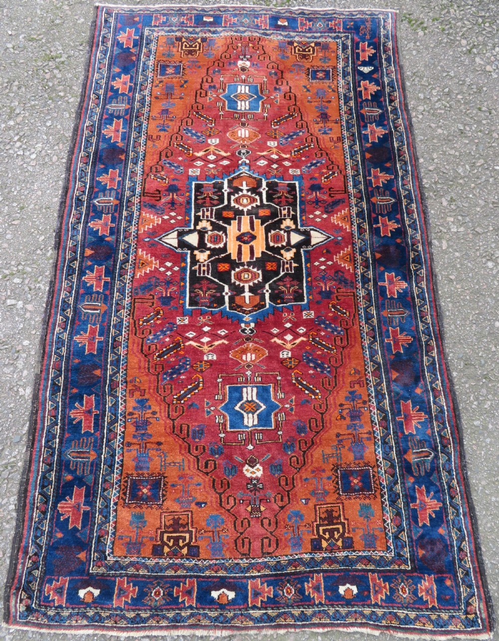 A hand-knotted Baluch wool rug featuring a central stepped dark medallion on a terracotta field bordered by geometric indigo patterns.