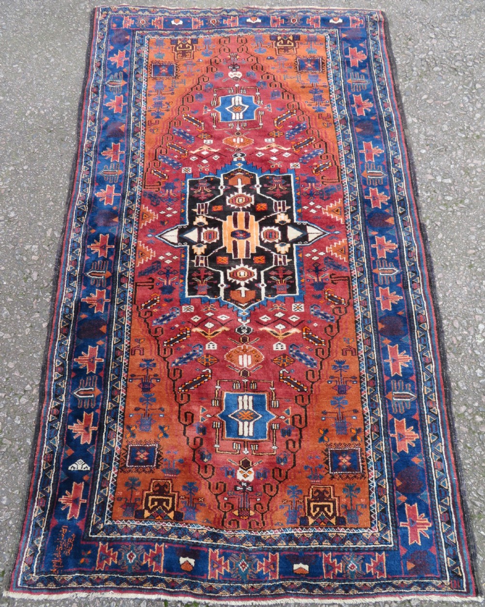 A hand-knotted Baluch wool rug featuring a central stepped dark medallion on a terracotta field bordered by geometric indigo patterns. View II