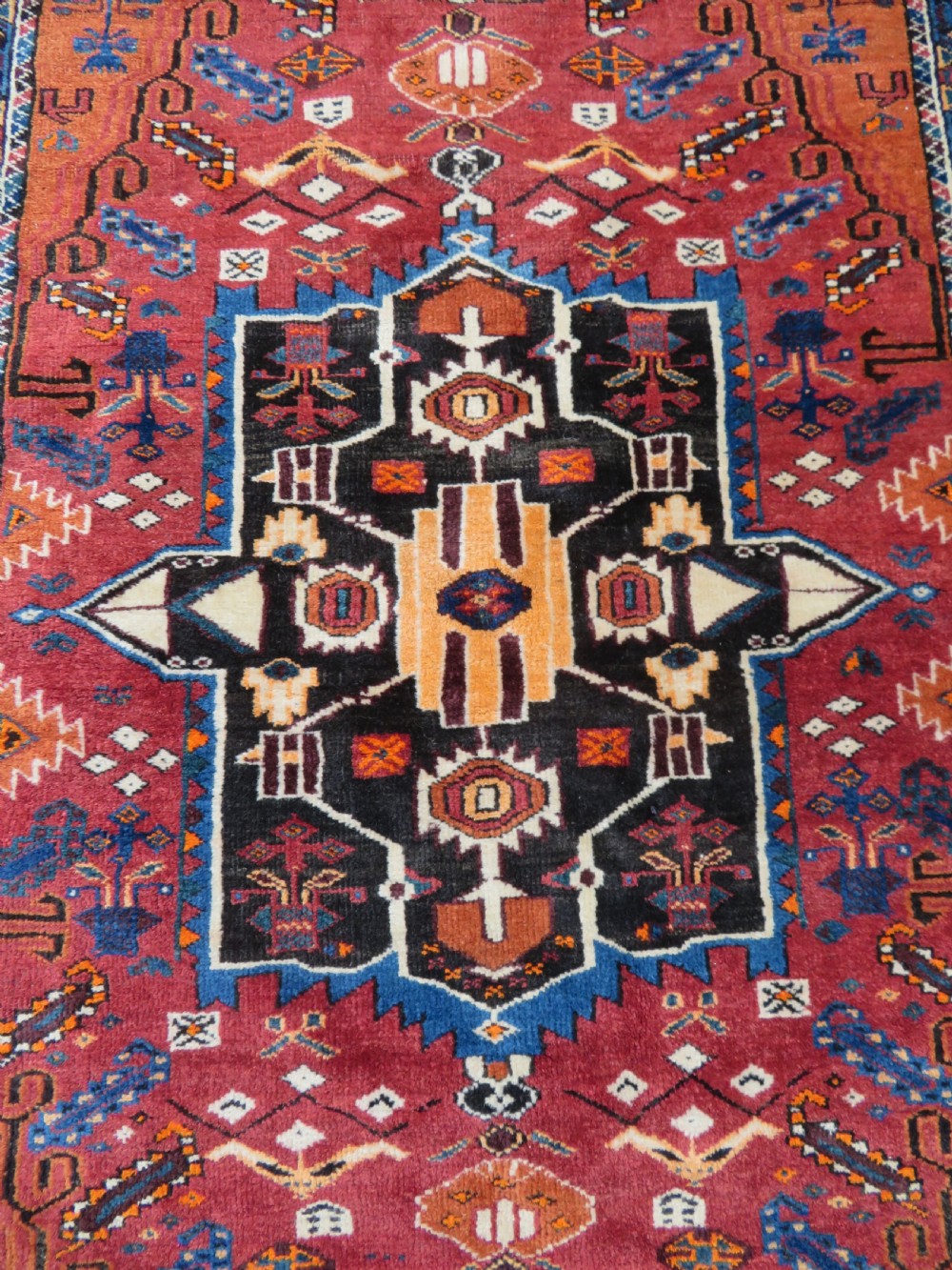 A hand-knotted Baluch wool rug featuring a central stepped dark medallion on a terracotta field bordered by geometric indigo patterns. View III