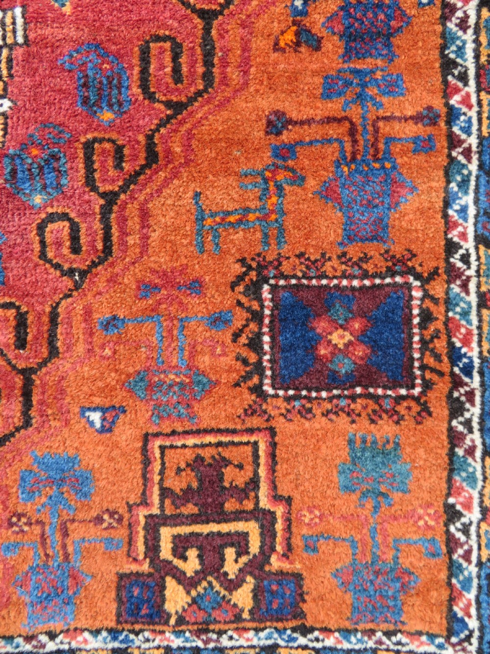 A hand-knotted Baluch wool rug featuring a central stepped dark medallion on a terracotta field bordered by geometric indigo patterns. View IV