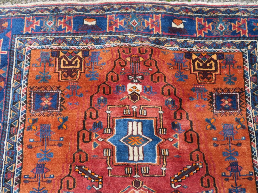 A hand-knotted Baluch wool rug featuring a central stepped dark medallion on a terracotta field bordered by geometric indigo patterns. View VI
