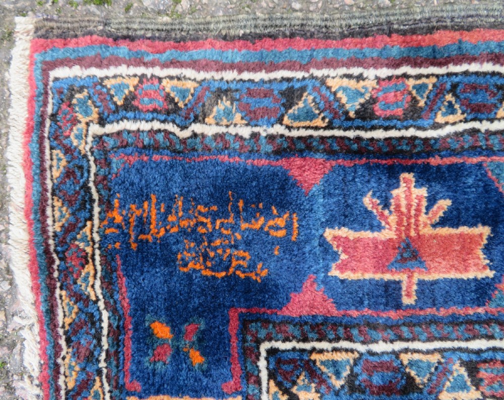 A hand-knotted Baluch wool rug featuring a central stepped dark medallion on a terracotta field bordered by geometric indigo patterns. View VII