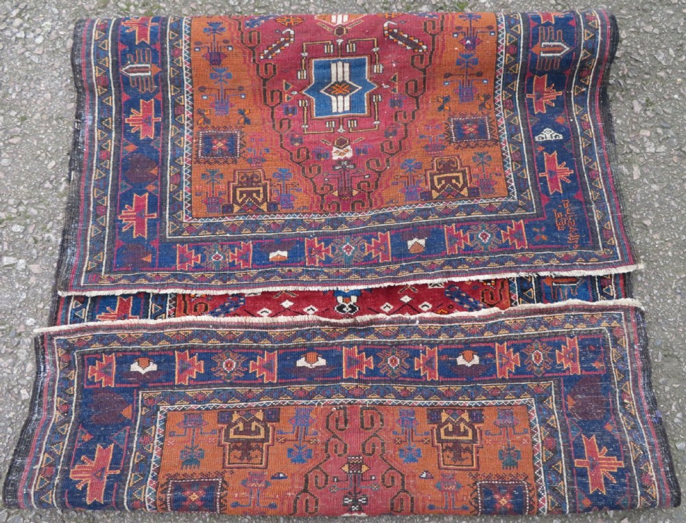 A hand-knotted Baluch wool rug featuring a central stepped dark medallion on a terracotta field bordered by geometric indigo patterns. View IX