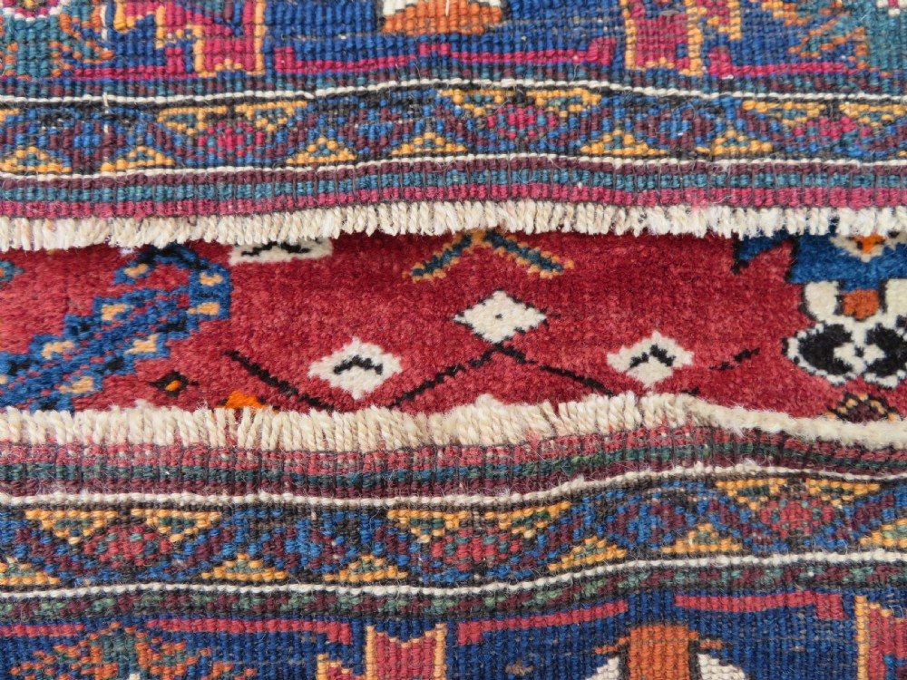 A hand-knotted Baluch wool rug featuring a central stepped dark medallion on a terracotta field bordered by geometric indigo patterns. View X