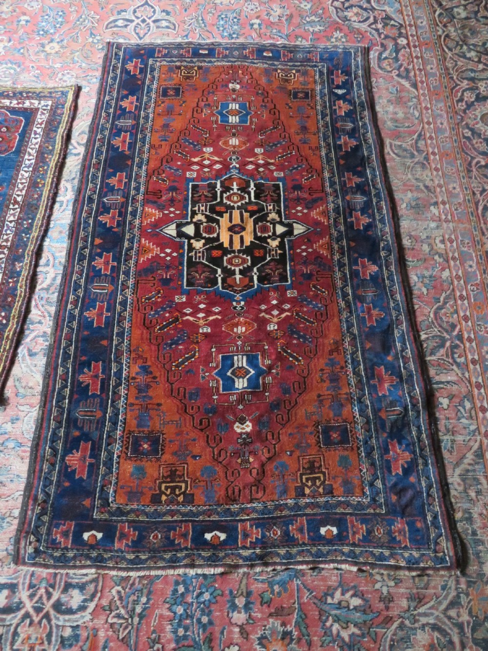 A hand-knotted Baluch wool rug featuring a central stepped dark medallion on a terracotta field bordered by geometric indigo patterns. View XII