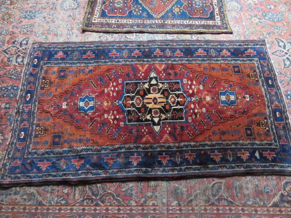 A hand-knotted Baluch wool rug featuring a central stepped dark medallion on a terracotta field bordered by geometric indigo patterns. View XIII