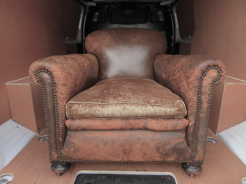 A distressed brown leather Art Deco club armchair with brass stud trimming along the rolled arms and a velvet seat cushion.