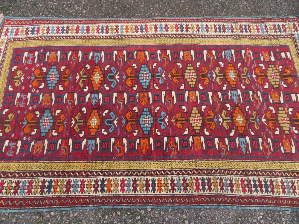 An antique hand-knotted Caucasian Karabagh wool rug featuring a red central field filled with repeating geometric motifs within multi-tiered yellow, blue, and cream borders. View II