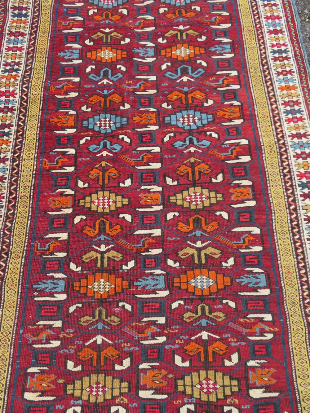 An antique hand-knotted Caucasian Karabagh wool rug featuring a red central field filled with repeating geometric motifs within multi-tiered yellow, blue, and cream borders. View III