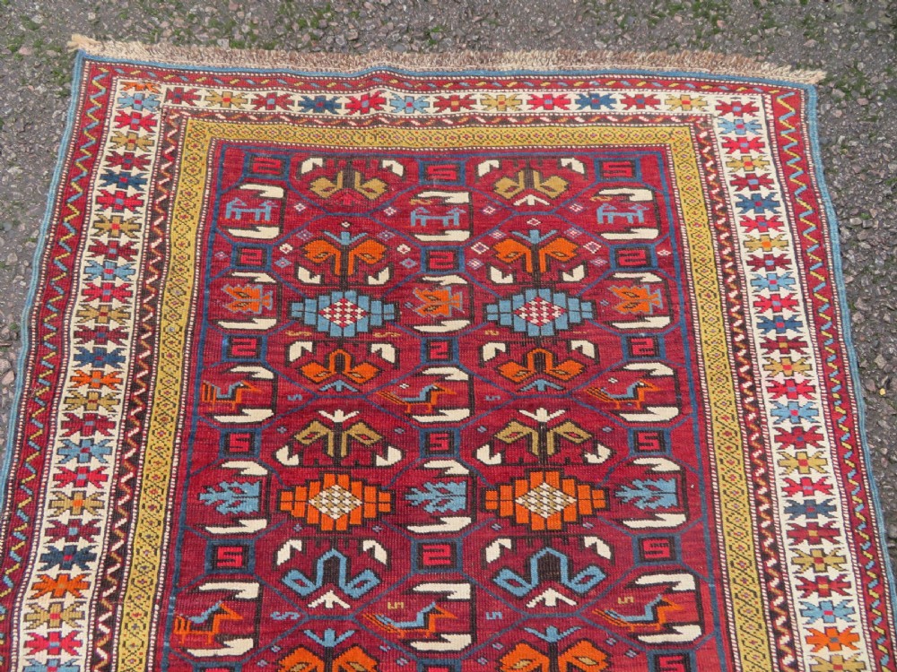 An antique hand-knotted Caucasian Karabagh wool rug featuring a red central field filled with repeating geometric motifs within multi-tiered yellow, blue, and cream borders. View IV
