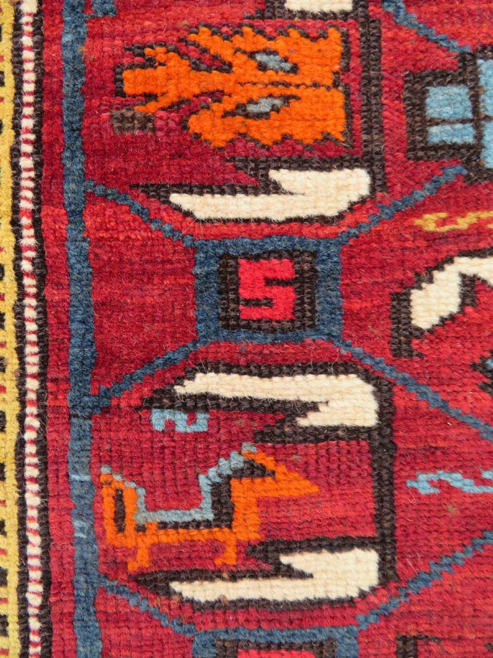 An antique hand-knotted Caucasian Karabagh wool rug featuring a red central field filled with repeating geometric motifs within multi-tiered yellow, blue, and cream borders. View V