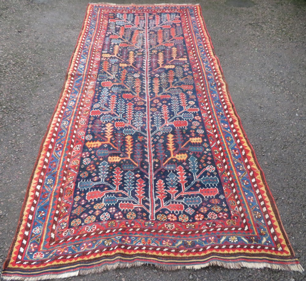 An antique hand-knotted wool kelleh rug featuring a central tree of life motif in red, yellow, and light blue against a dark navy field.