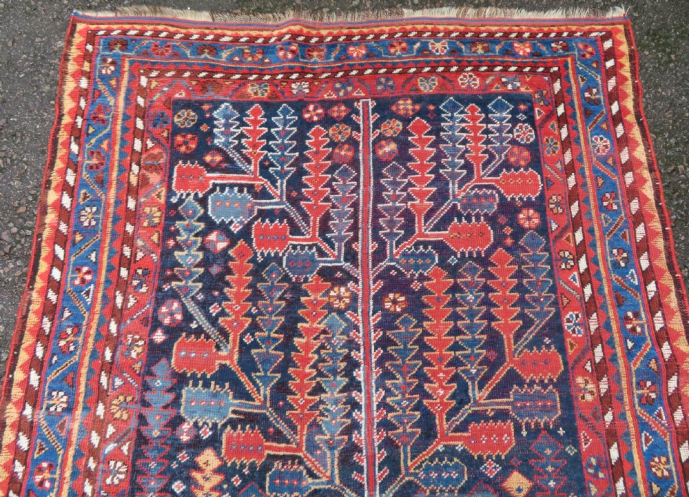 An antique hand-knotted wool kelleh rug featuring a central tree of life motif in red, yellow, and light blue against a dark navy field. View II