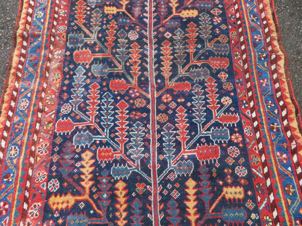 An antique hand-knotted wool kelleh rug featuring a central tree of life motif in red, yellow, and light blue against a dark navy field. View IV