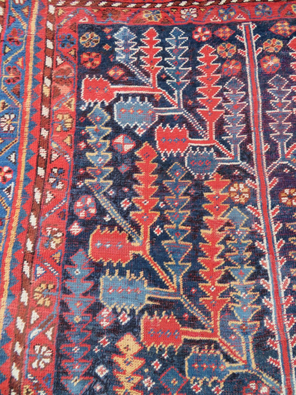 An antique hand-knotted wool kelleh rug featuring a central tree of life motif in red, yellow, and light blue against a dark navy field. View X
